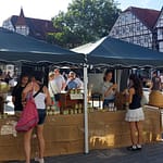 175 Jahre Leidenschaft: Wie der Imkerverein Soest zwischen Tradition und Hornissen-Invasion begeistert 17 Zwei junge Frauen stehen vor einem Marktstand mit Honiggläsern auf einem gepflasterten Marktplatz, im Hintergrund historische Fachwerkhäuser und eine Baumreihe auf dem Soester Bauernmarkt.