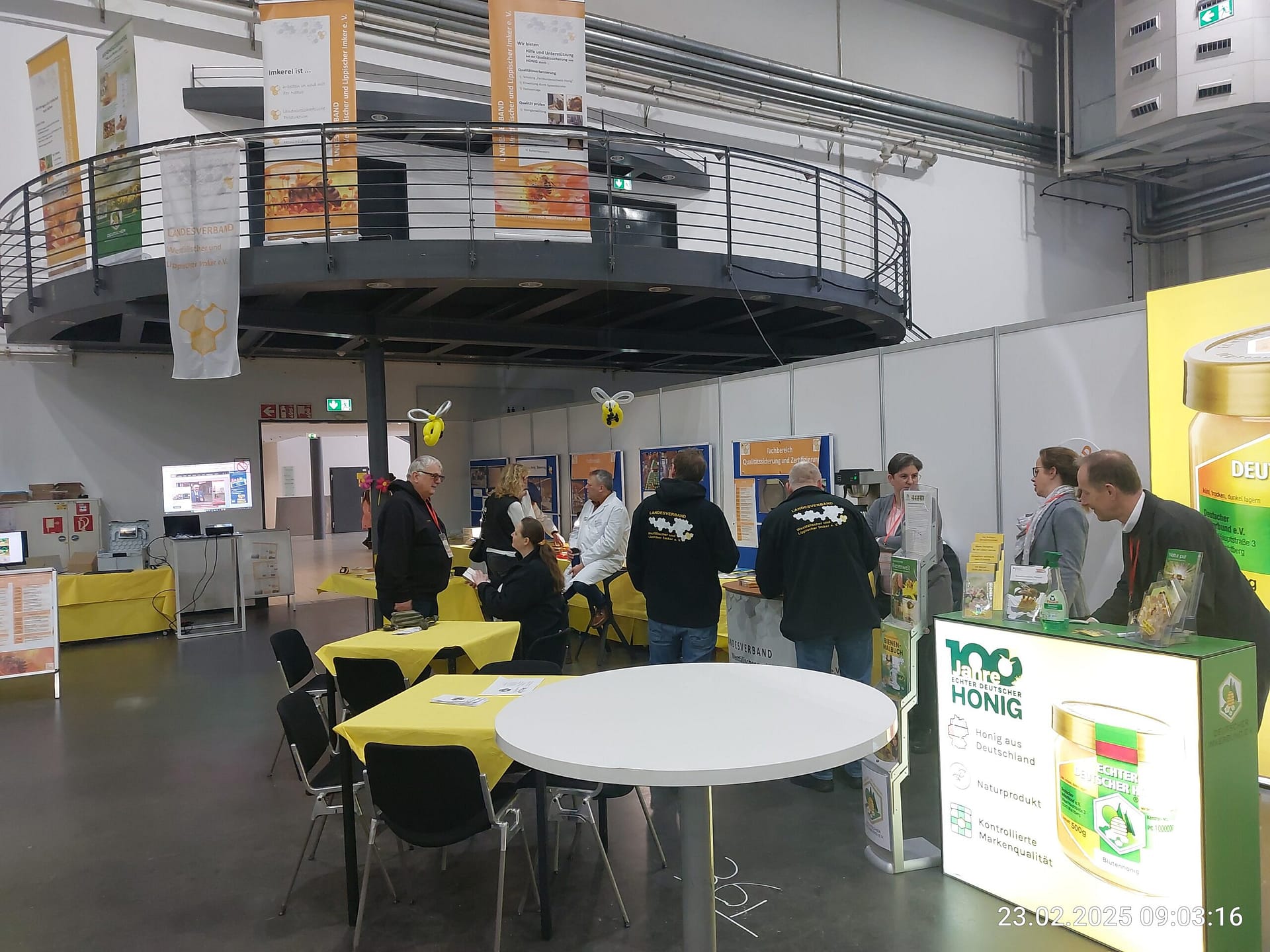 Einblick in einen gut besuchten Messestand auf der "Echter Deutscher Honig"-Ausstellung, mit Besuchern, die sich an Tischen unterhalten und Informationsstände betrachten, darunter ein Stand mit der Aufschrift "100 Jahre Echter Deutscher Honig".