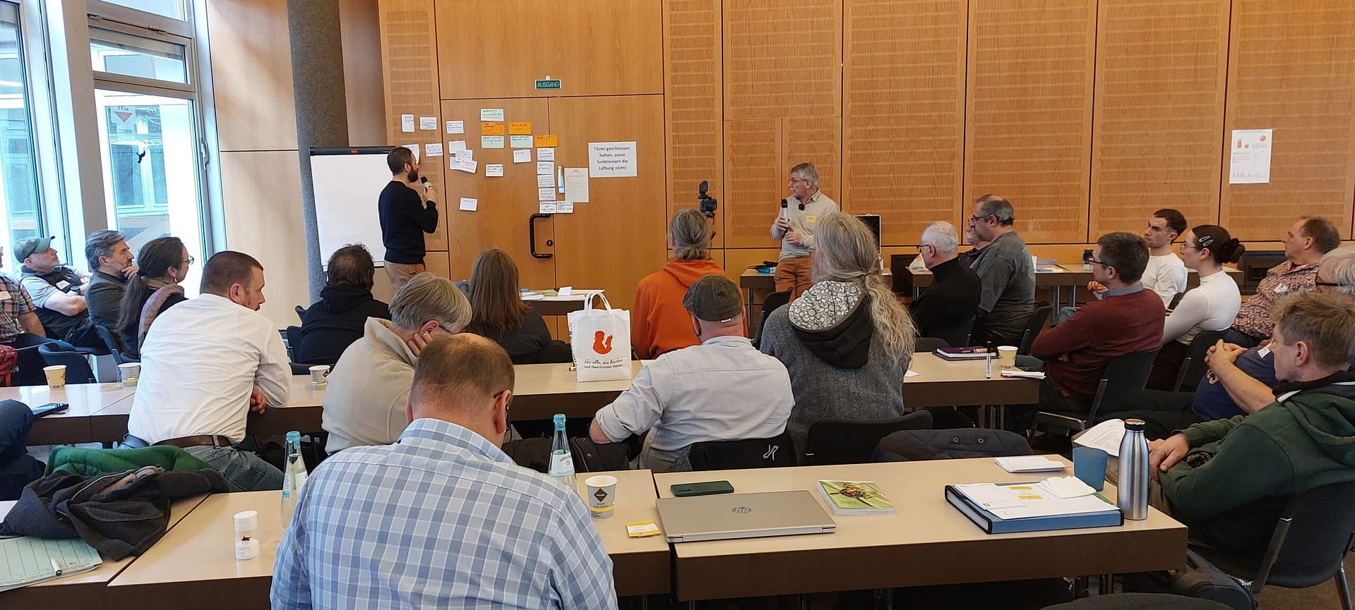 Ein Moderator präsentiert an einer mit Post-its beklebten Holzwand vor einer Gruppe von Teilnehmern in einem Seminarraum.