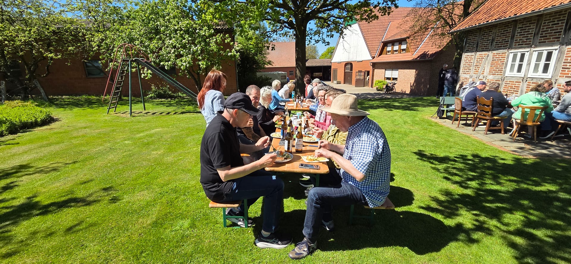 Imkerverein Herford | Bienenhaltung, Imkerei, Honig & Ausbildung 29 Mitglieder des Imkervereins Herford sitzen bei sonnigem Wetter an Biertischen auf einer grünen Wiese vor einem historischen Fachwerk-Bauernhof und essen gemeinsam.
