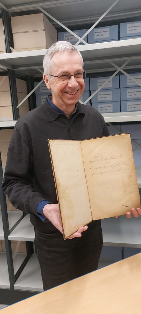 Von Sütterlin bis Cloud: Herforder Imker retten 140 Jahre Bienengeschichte 2 Udo Rolfsmeier im Archiv hält ein aufgeschlagenes, historisches Protokollbuch in die Kamera. Im Hintergrund sind Regale mit Archivkartons des Kreisarchivs Herford zu sehen.